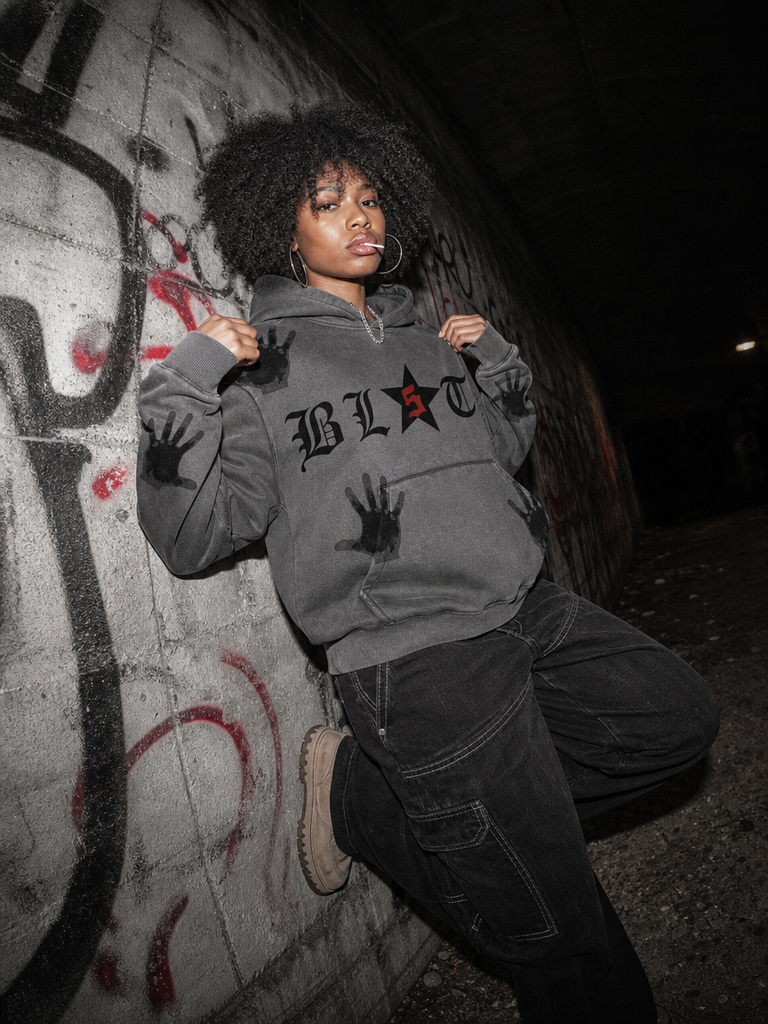 A woman with an afro hairstyle poses against a graffiti-covered concrete wall, wearing a dark gray hoodie with 'BL5TT' and a star graphic on the chest, black cargo pants, and beige sneakers.